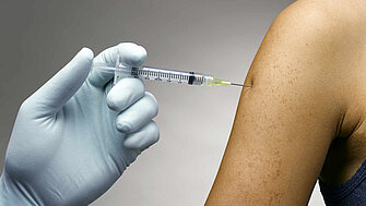 Behandschuhte Hand mit Impfspritze verabreicht eine Impfung in einen weiblichen Oberarm.